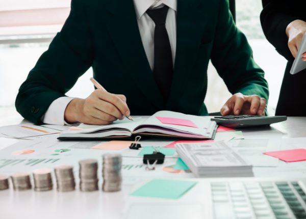 Eine Person sitzt an einem Tisch mit einem Taschenrechner, Notizbuch und Zetteln. Ein Stapel Geld liegt im Vordergrund