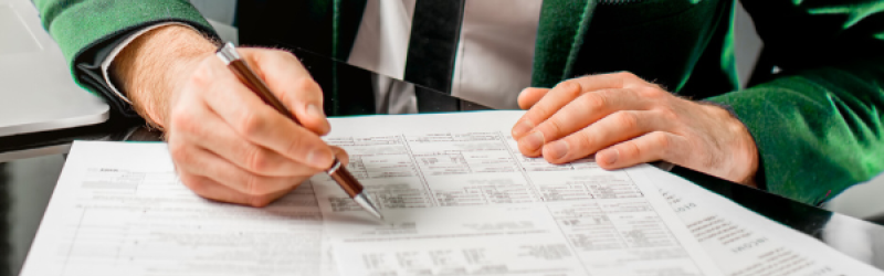 Eine Person in grünem Anzug, die ein Kompliziert aussehendes Formular an einem Tisch ausfüllt.